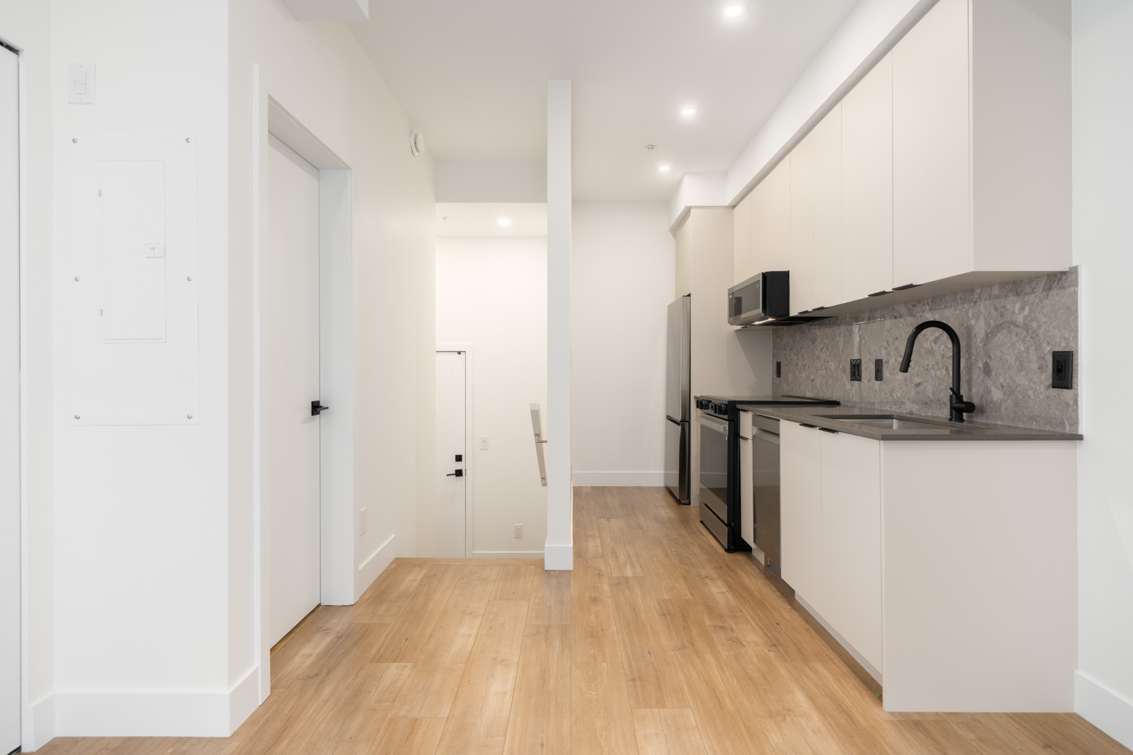 Modern kitchen with light wood flooring, white cabinets, stainless steel appliances, and a gray backsplash; stairs and doors visible in the background.