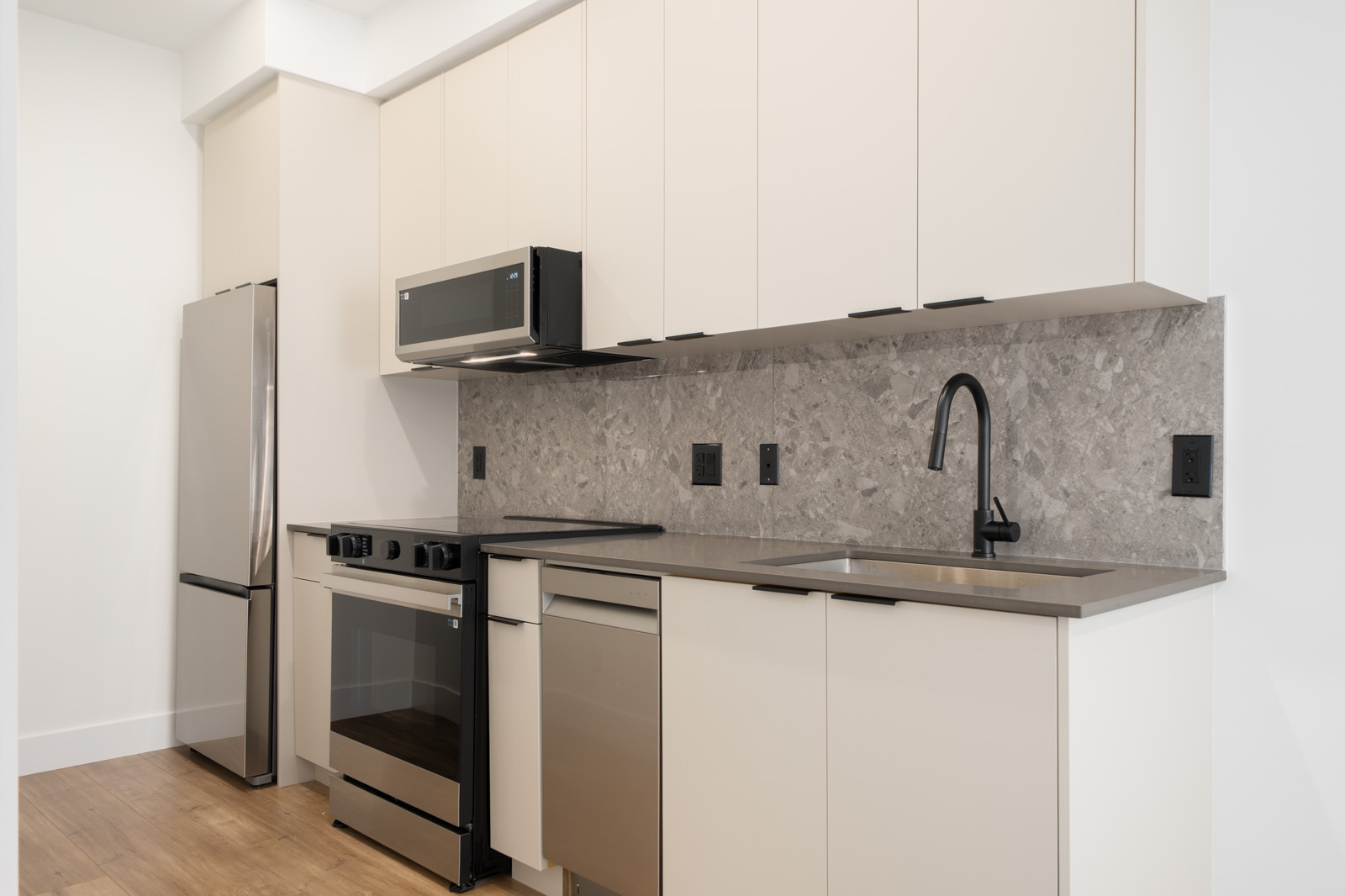 Modern kitchen with stainless steel appliances, beige cabinets, gray stone backsplash, black fixtures, and light wood flooring.