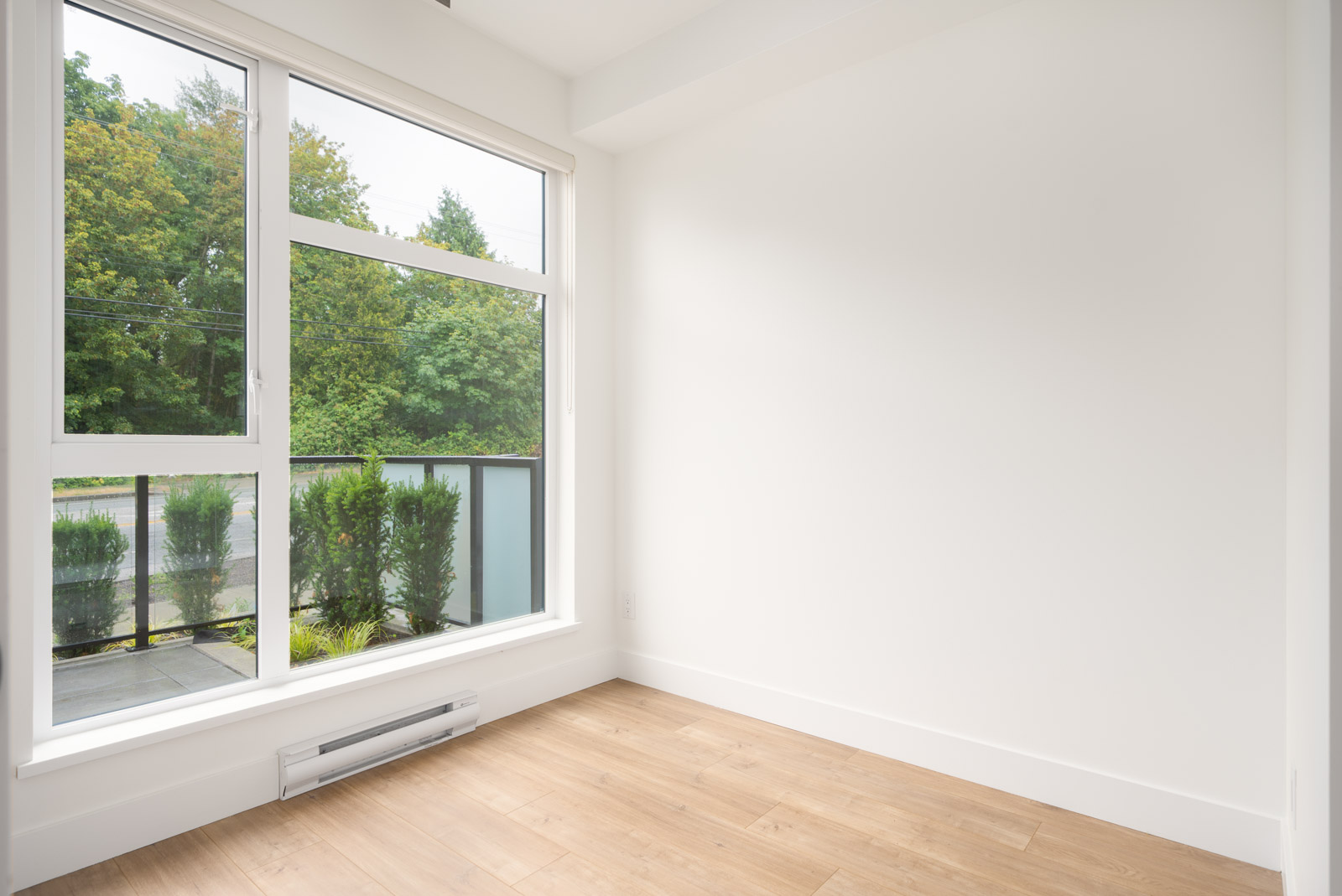 Empty room with white walls, large window overlooking greenery, light wood flooring, and a baseboard heater under the window.