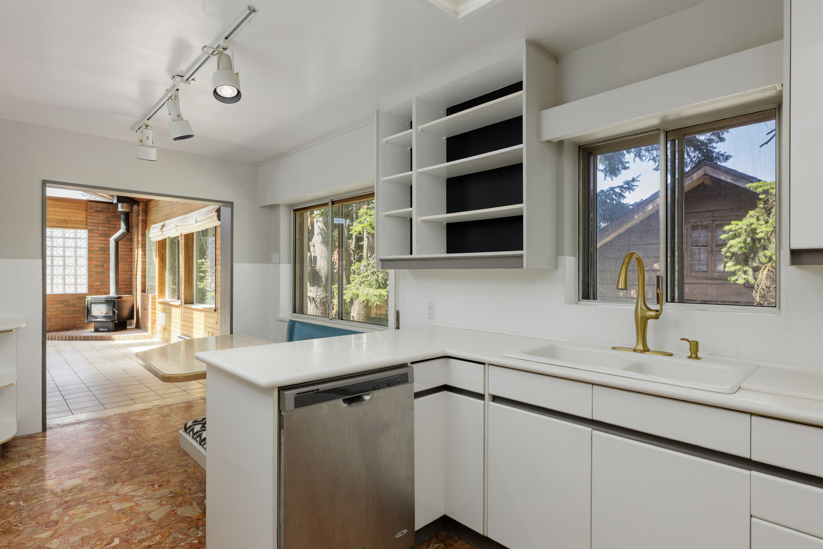 Modern kitchen with white cabinets, open shelves, gold faucet, stainless dishwasher, and large windows next to sunroom in Burnaby.