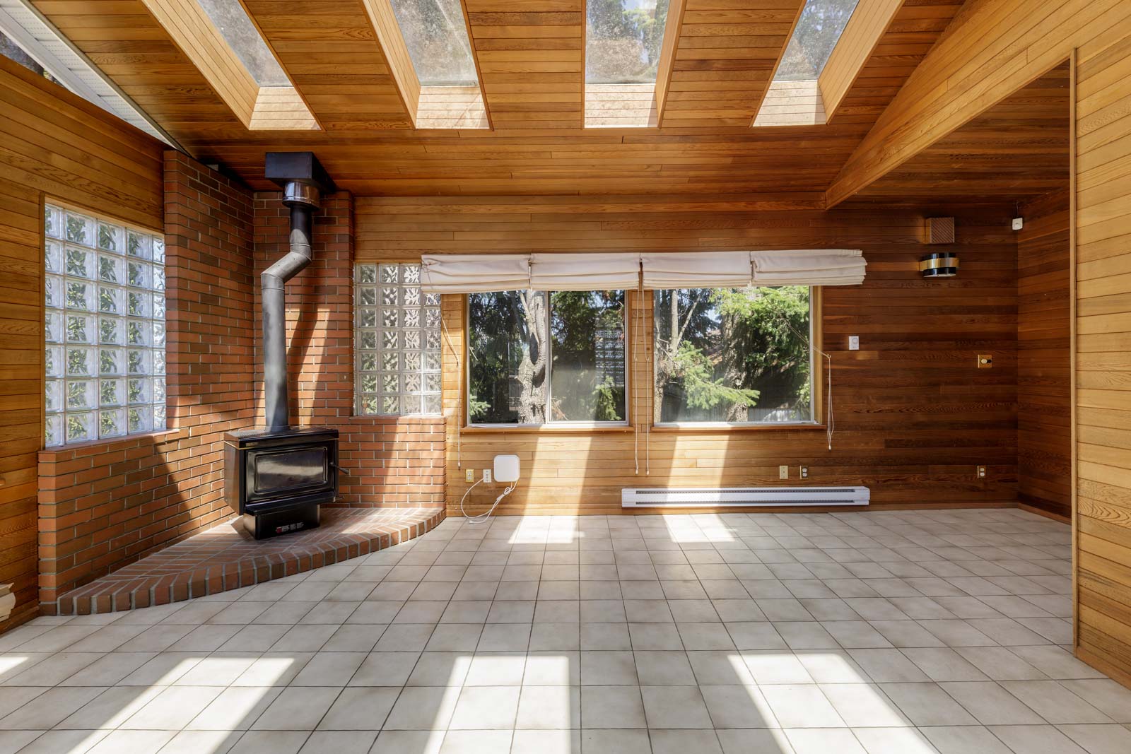 Bright Burnaby room with wood-paneled walls, tiled floor, skylights, large windows, brick fireplace stove, and baseboard heater.