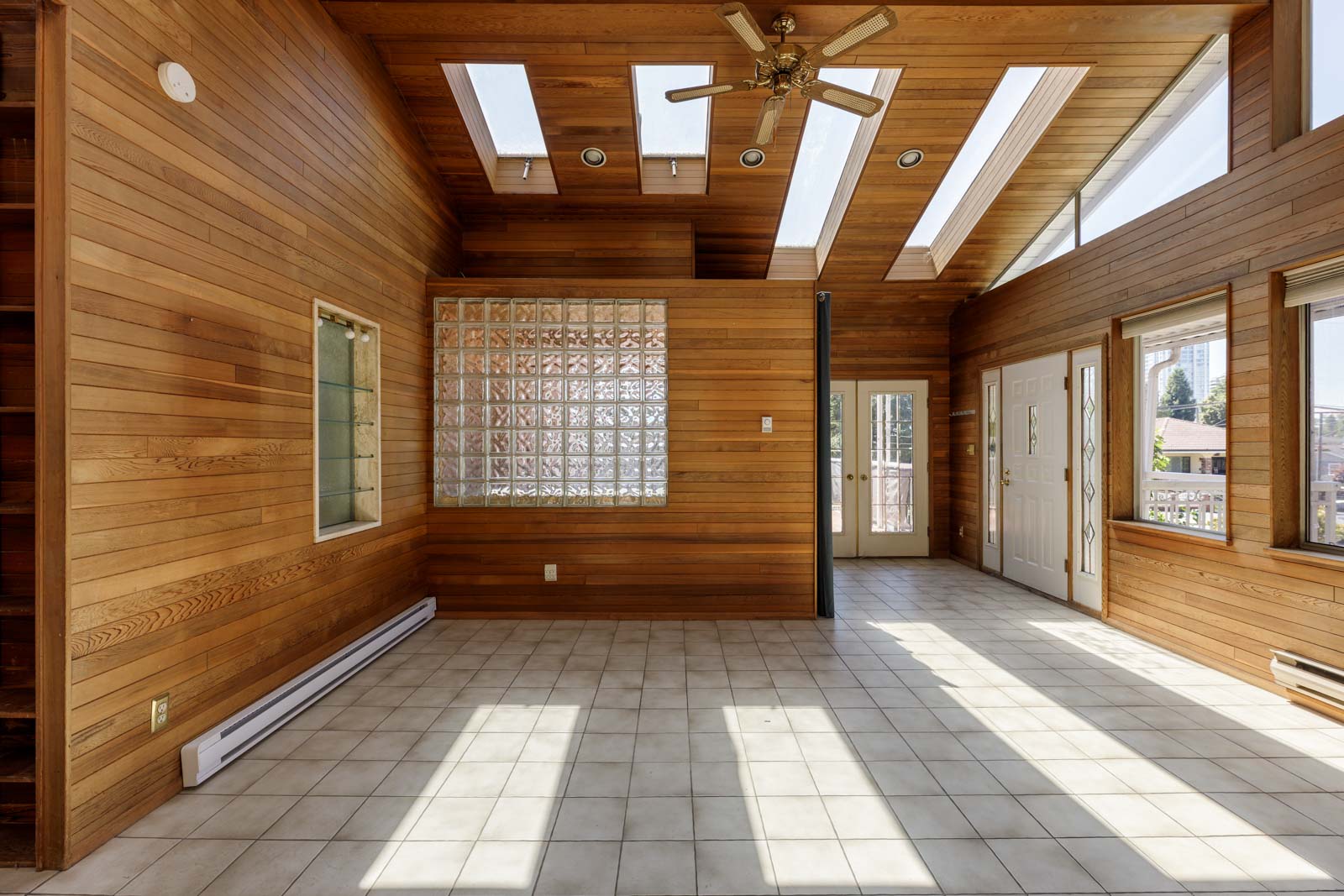 Room at 6791 Griffiths Ave, Burnaby with wood-paneled walls, tiled floor, skylights, glass block divider, fan, and large windows.