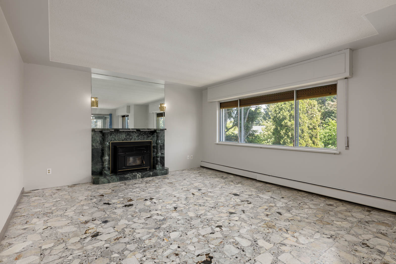 Empty living room with marble-patterned floor, large window, black fireplace in a mirrored wall, and sconces in Burnaby.