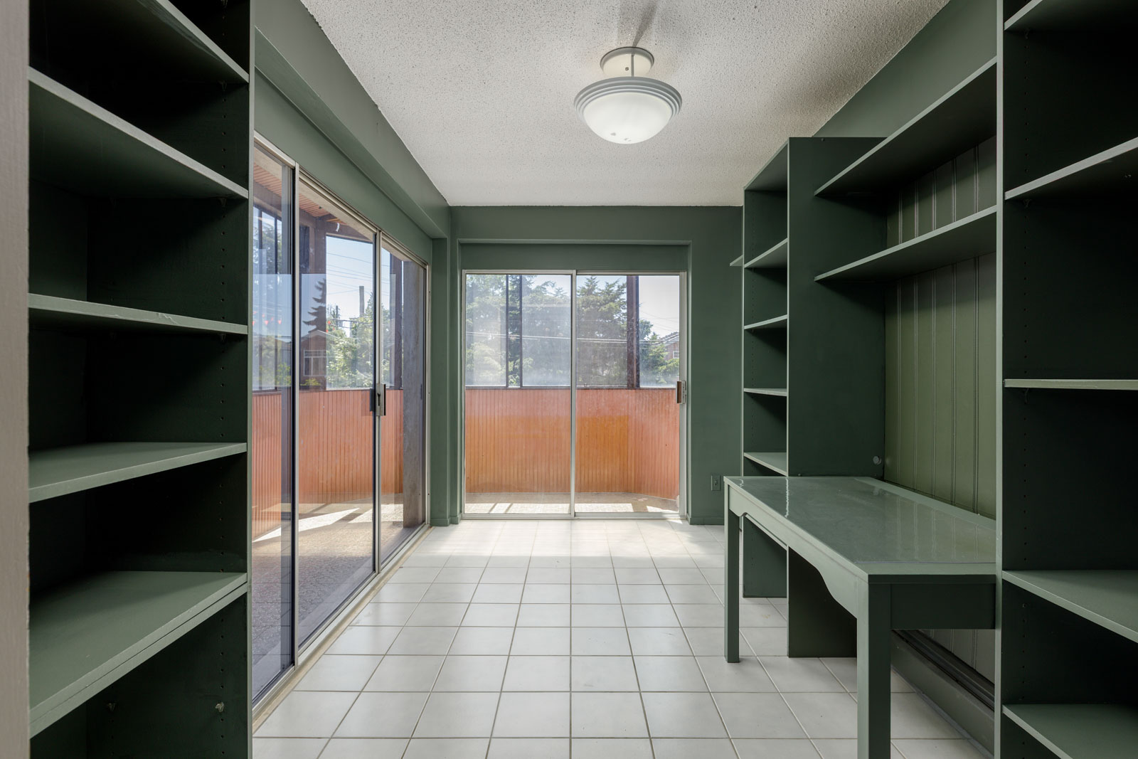 Room with built-in green shelves and desk, white tile flooring, and sliding glass doors to a balcony on Upper Griffiths Ave, Burnaby.