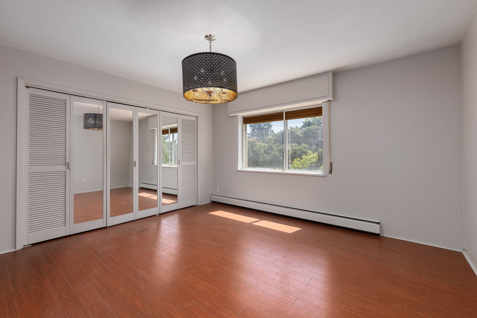 Bedroom at 6791 Griffiths Ave in Burnaby with light brown wood flooring, gray walls, mirrored closets, window, and black chandelier.