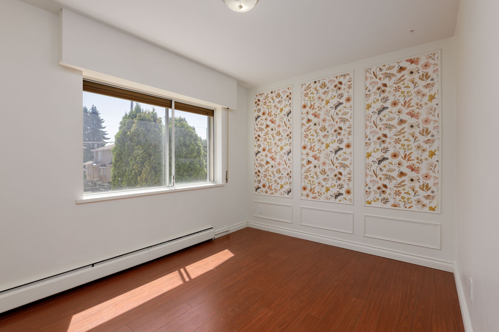 Empty room with wood floors, large window, and three floral wallpaper panels. Trees visible outside on Griffiths Ave, Upper Burnaby.