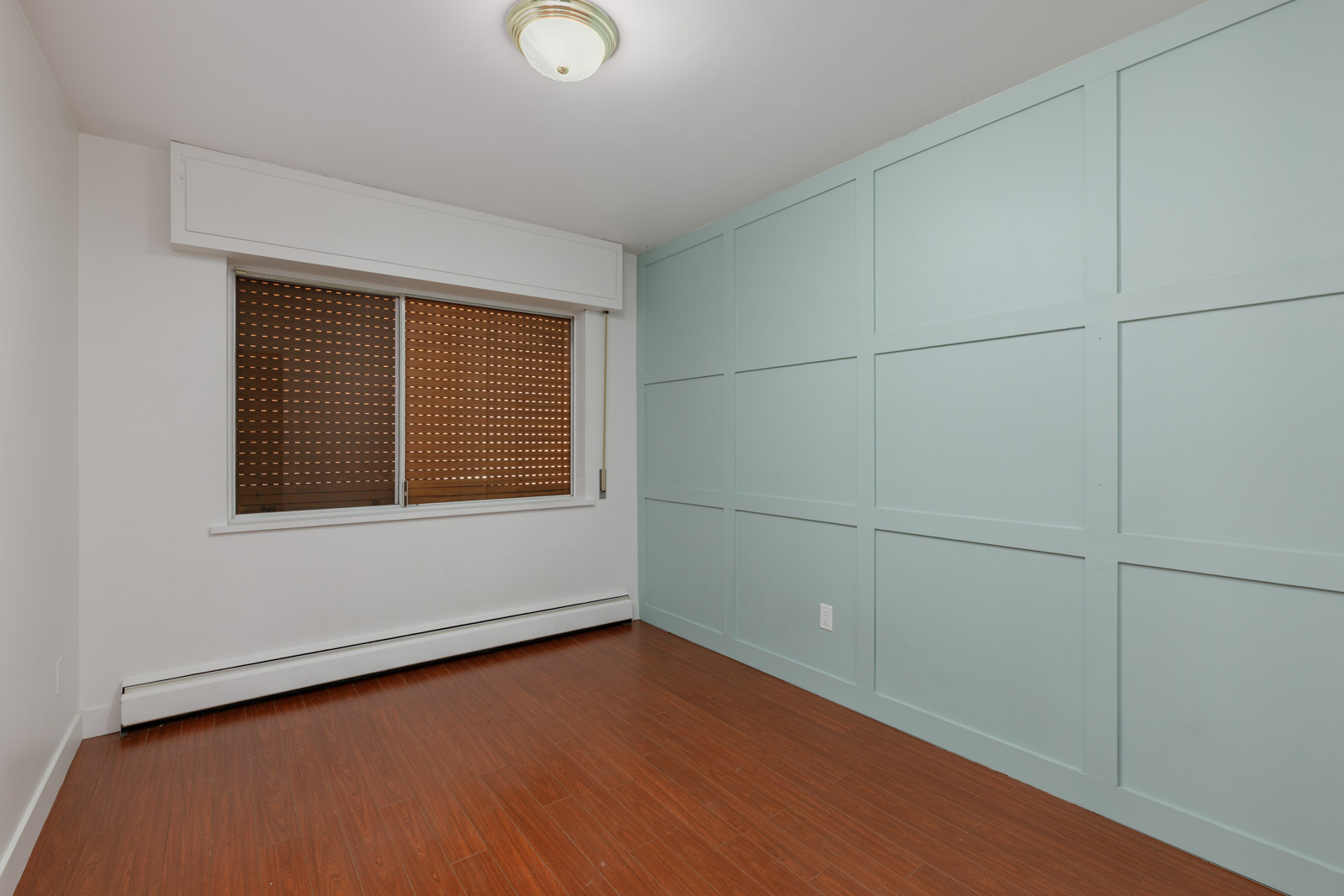 Empty upstairs room on Griffiths Ave, Burnaby with light blue paneled wall, wood floor, baseboard heater, window blinds, and ceiling light.
