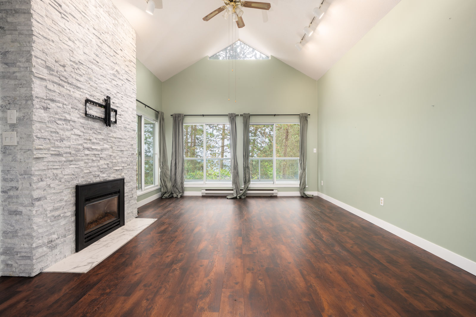 Living room with high ceiling, wood floors, stone fireplace, large windows with gray curtains, and a ceiling fan.