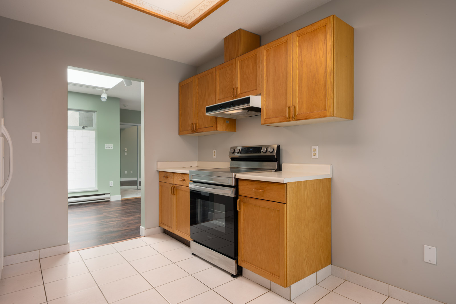 Kitchen featuring light wood cabinets, electric stove, white countertops, beige tile floor, and a view into a room with wood floors.