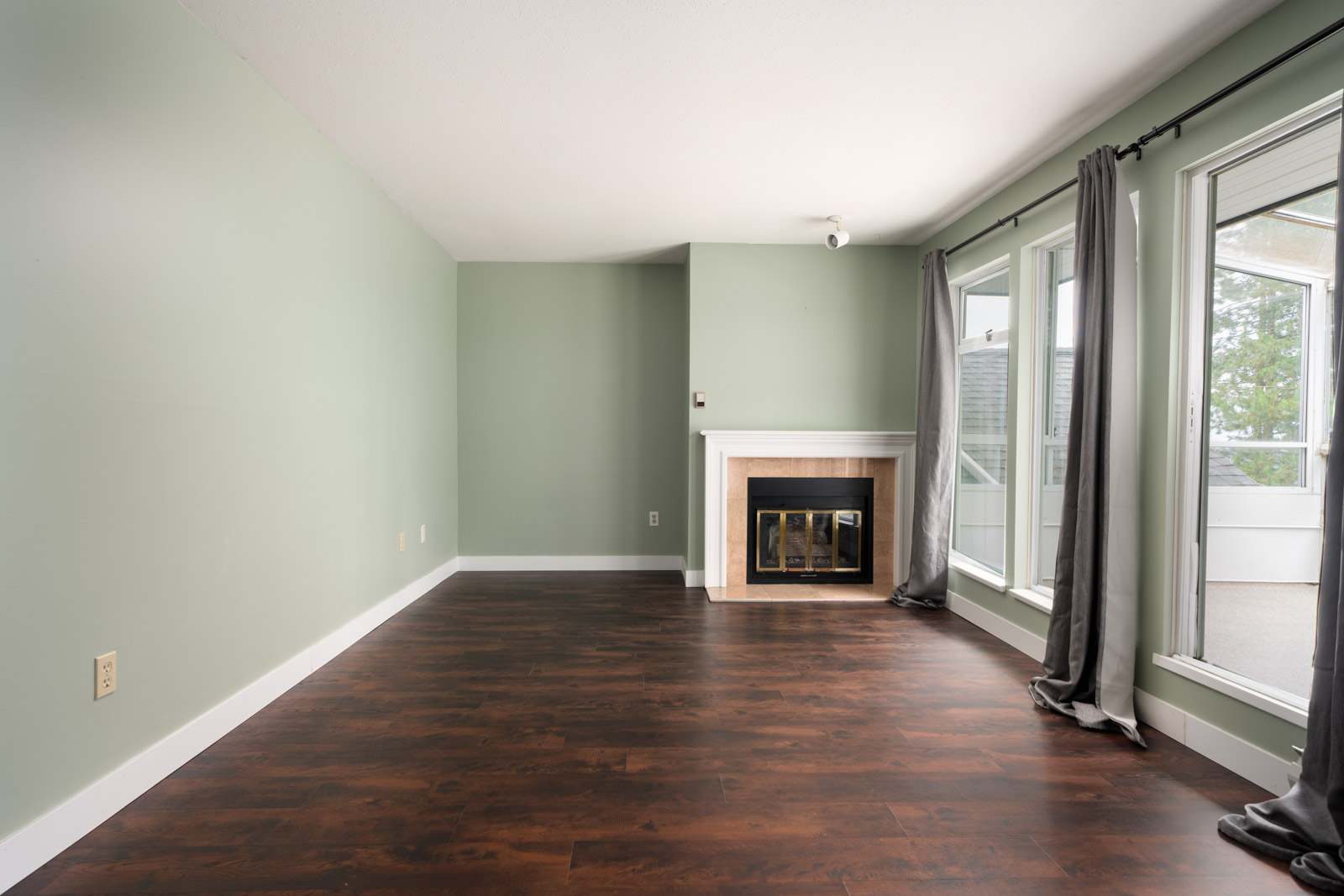 Empty living room with light green walls, dark wood floors, white fireplace, large windows with gray curtains, and balcony door.