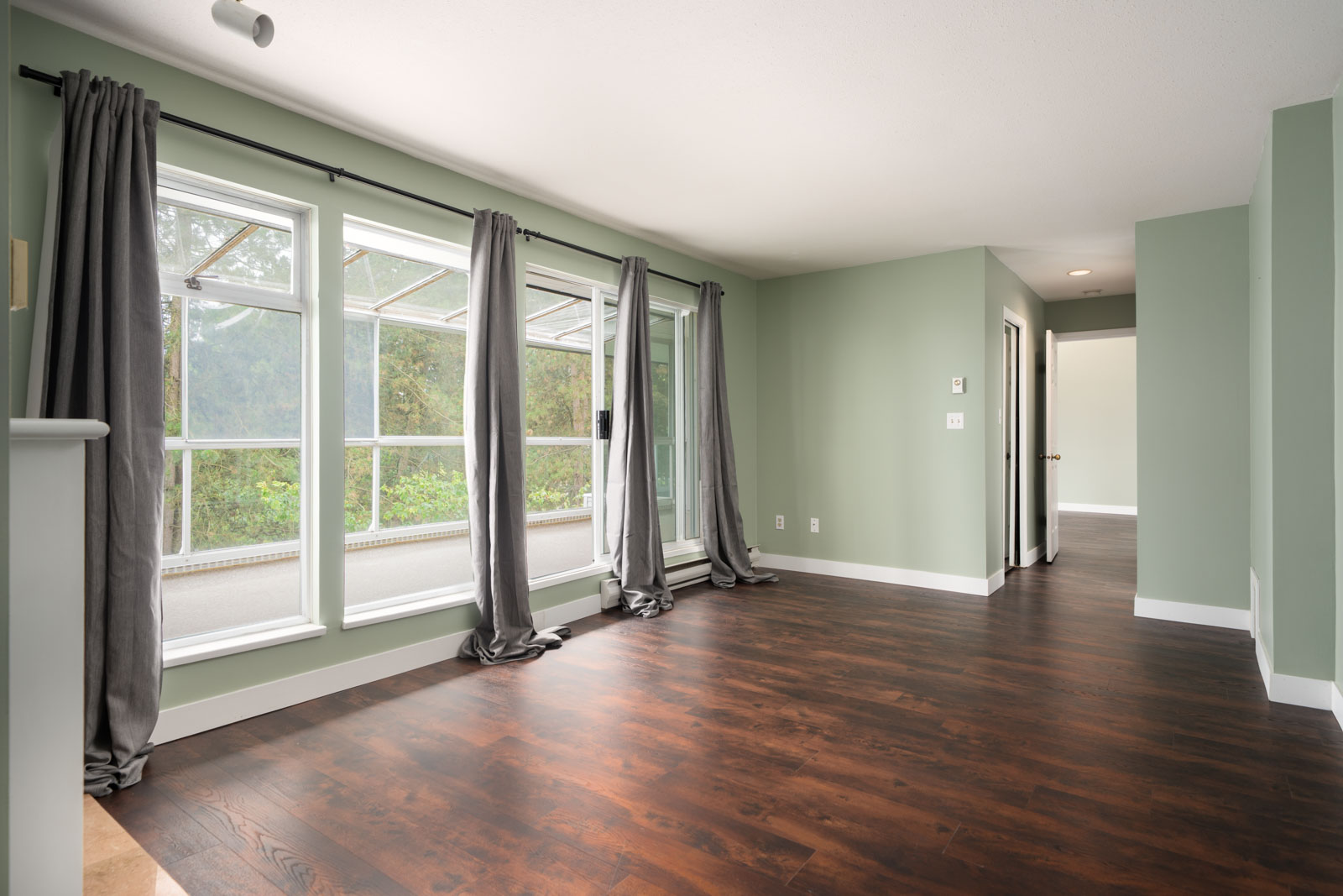Empty room with light green walls, dark wood floors, large windows, gray curtains, and a view of a balcony and trees.