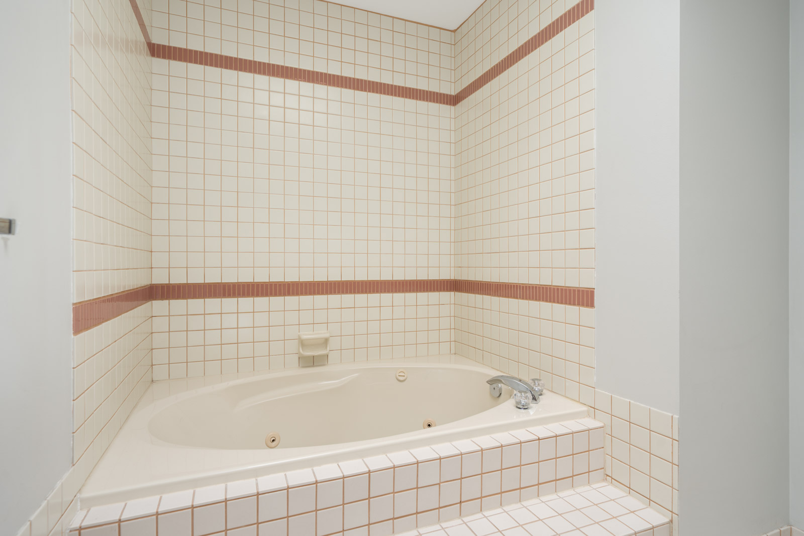 Cream-colored built-in bathtub with white tile surround, a pink accent row, wall-mounted soap holder, and chrome faucet.