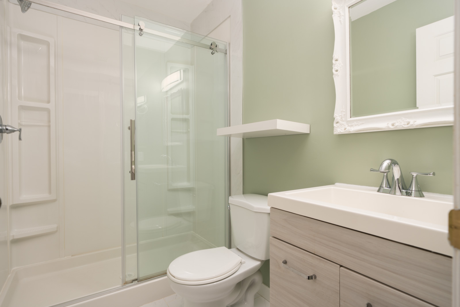 Bathroom featuring a glass shower, toilet, vanity with sink, wall shelf, and large mirror on a light green wall.