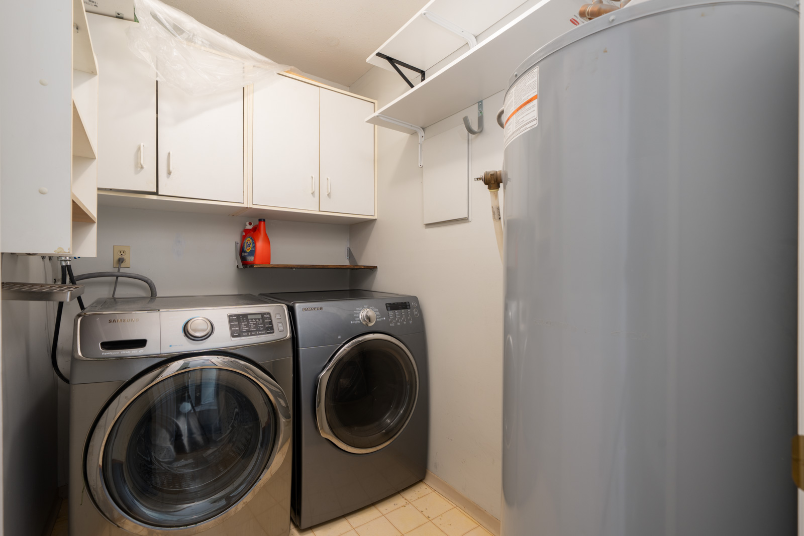 Laundry room featuring a front-loading washer and dryer, white cabinets, shelves, water heater, and cleaning supplies on a shelf.