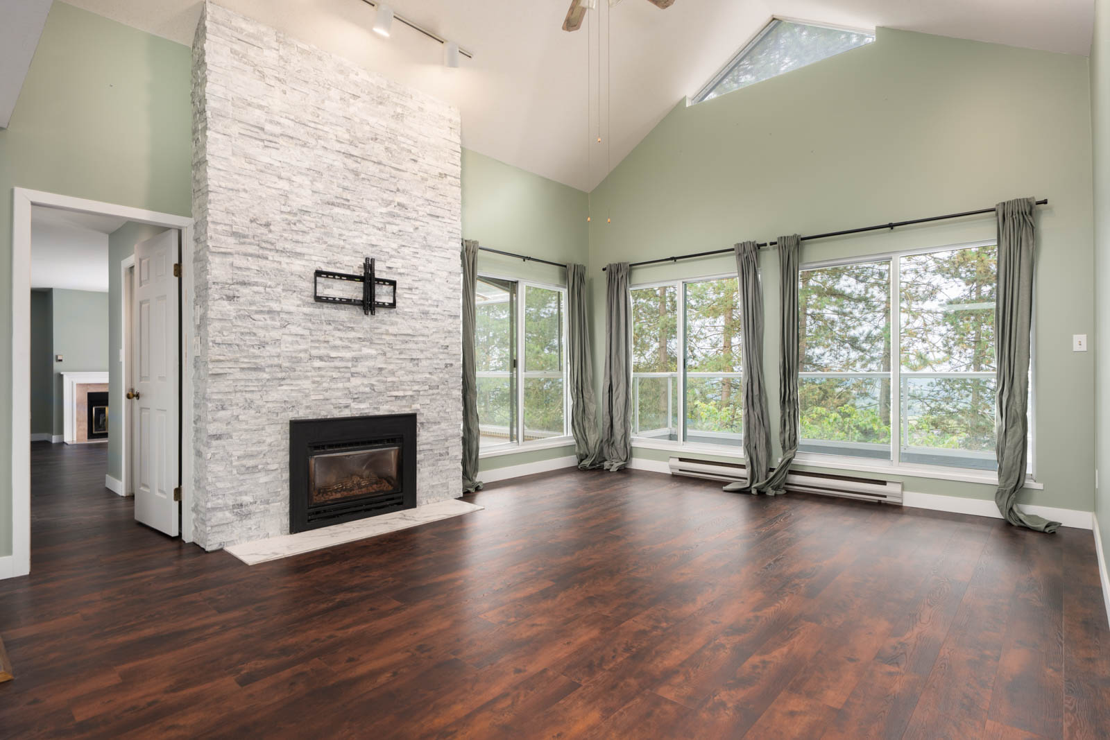 Spacious living room with large windows, stone fireplace, wood floors, vaulted ceiling, green walls, and gray curtains.