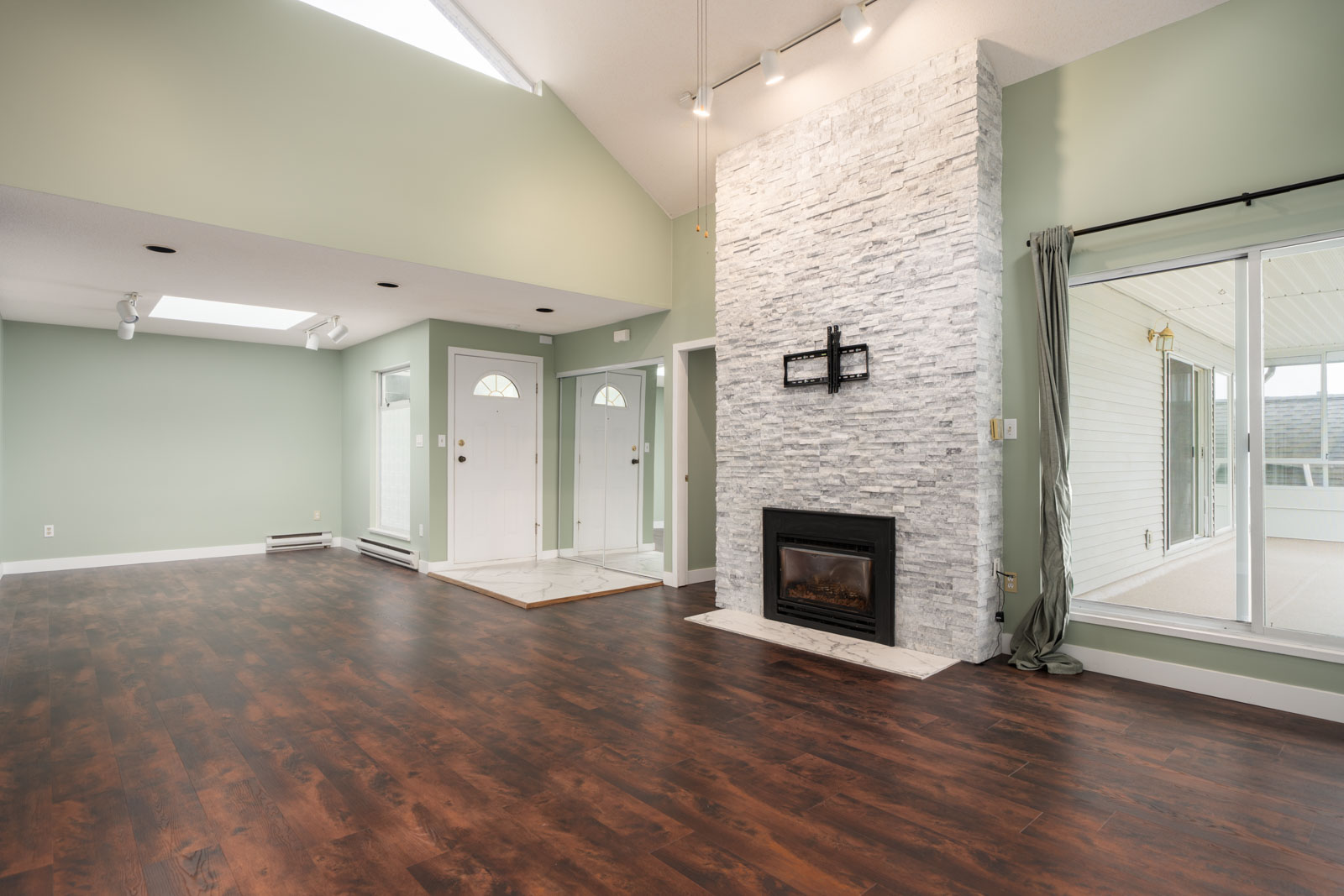 Living room with light green walls, vaulted ceiling, dark wood floors, stone fireplace, skylights, and glass doors to sunroom.