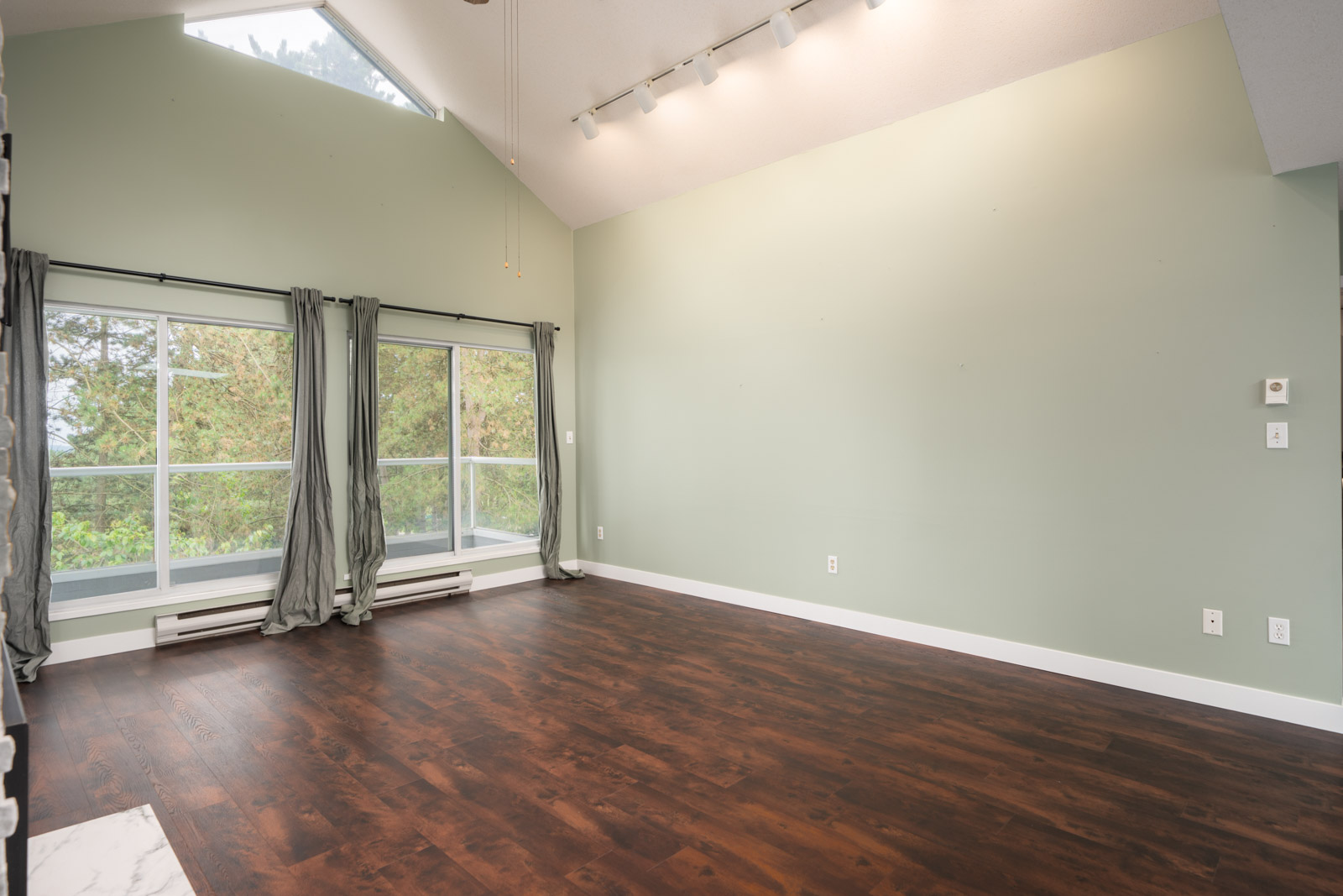 Spacious empty room with light green walls, dark wood floors, large windows with gray curtains, white trim, and track lighting.