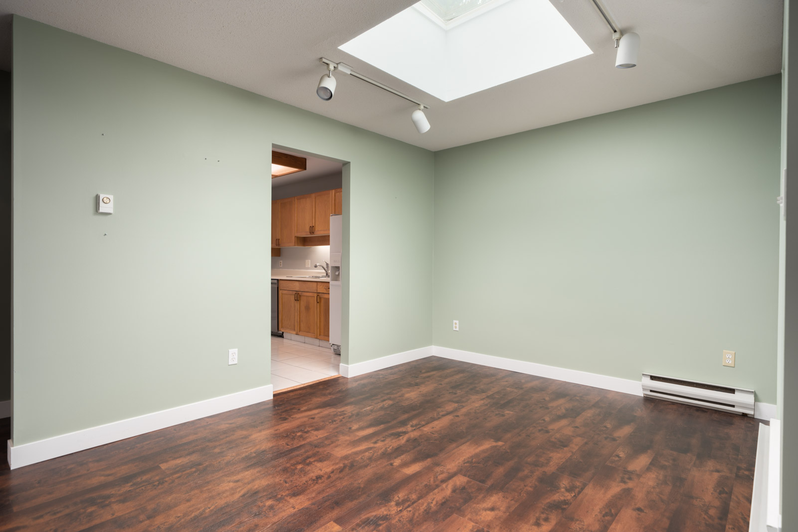 Empty room with green walls, dark wood floor, skylight, baseboard heater, and doorway to kitchen with wood cabinets and white tile.