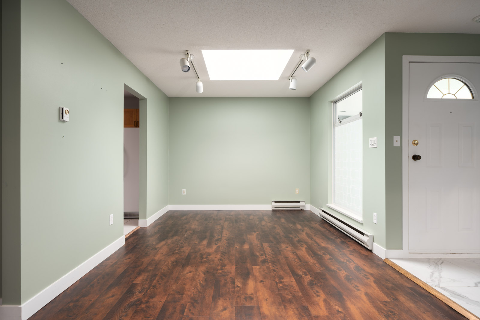 Empty room with mint green walls, dark wood floors, a white door, skylight, track lights, baseboard heater, and right-side window.