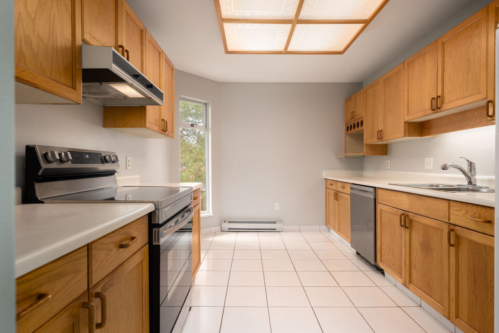 Contemporary kitchen featuring wooden cabinets, stainless appliances, white counters, light tile flooring, and a large sunlit window.