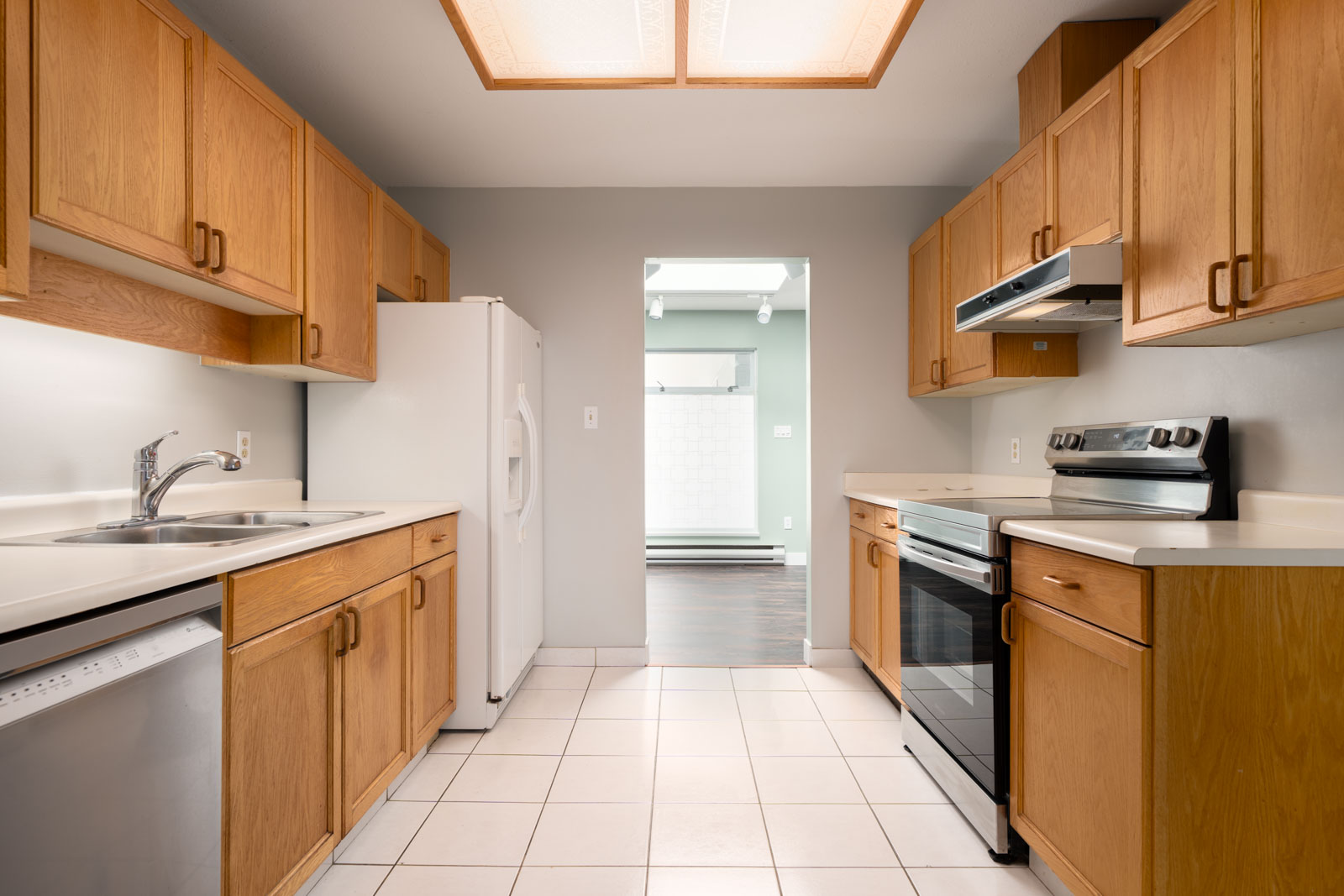 Galley kitchen with light wood cabinets, white appliances, electric stove, dishwasher, and white tile floor; doorway to hardwood room.