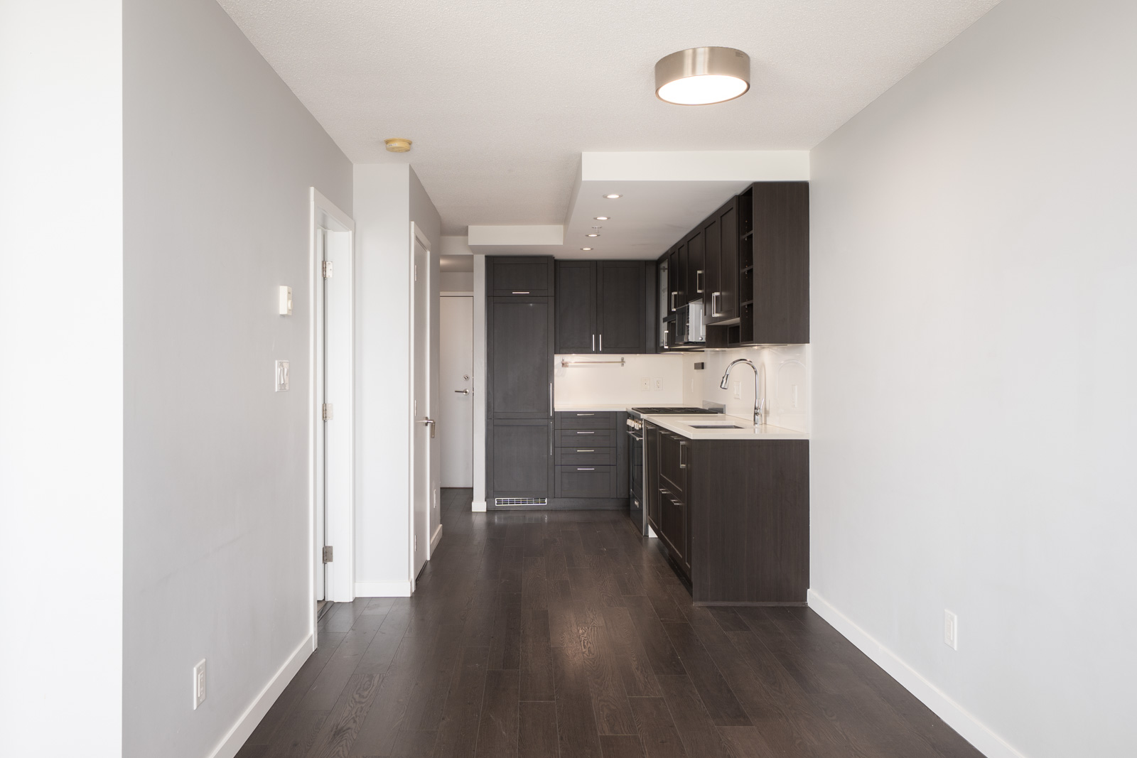 Empty modern Vancouver apartment kitchen with dark cabinets, white counters, wood floors, and hallway with white walls and doors.