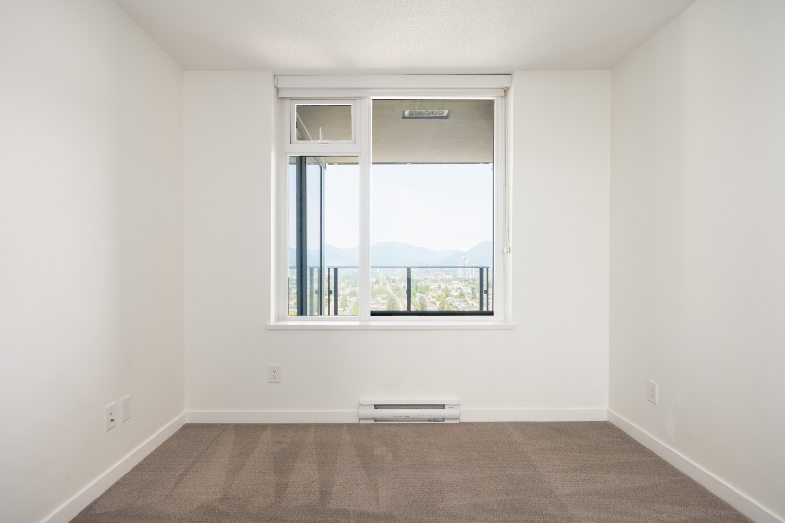 Empty room with beige carpet, white walls, and a large window overlooking a balcony with city and mountain views in Vancouver.