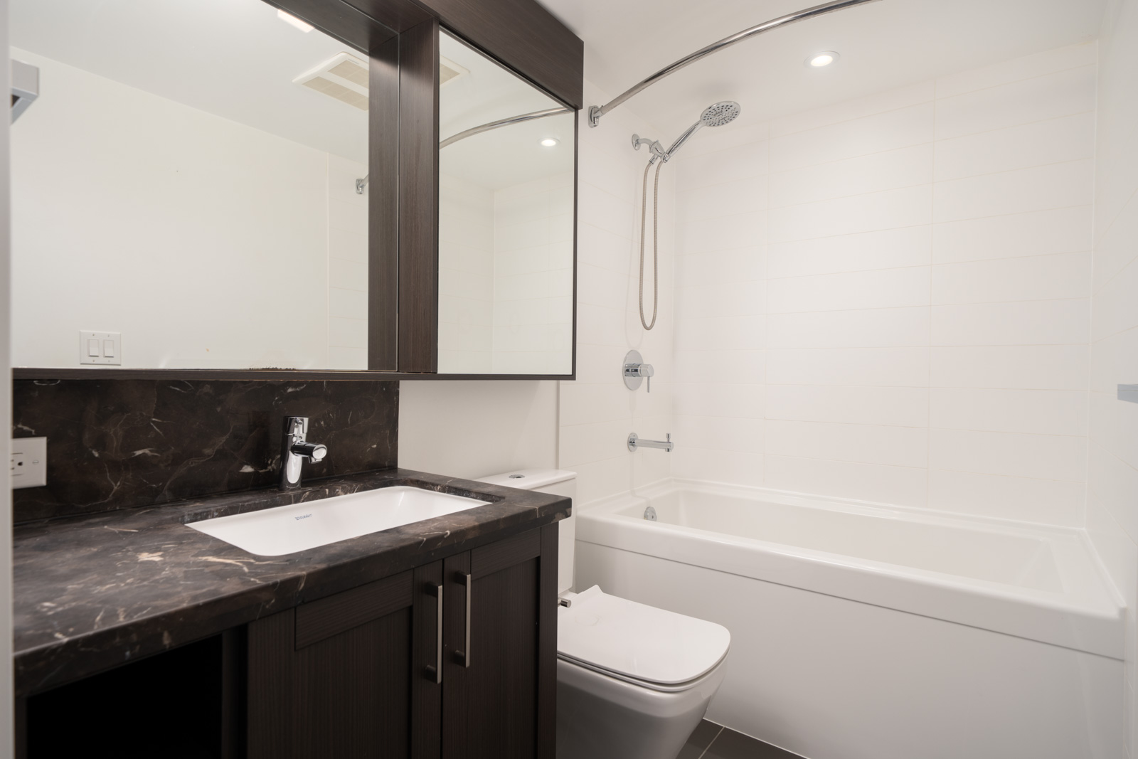 Modern Vancouver bathroom with dark wood vanity, marble counter, sink, mirror cabinet, and white tub with shower on tiled wall.