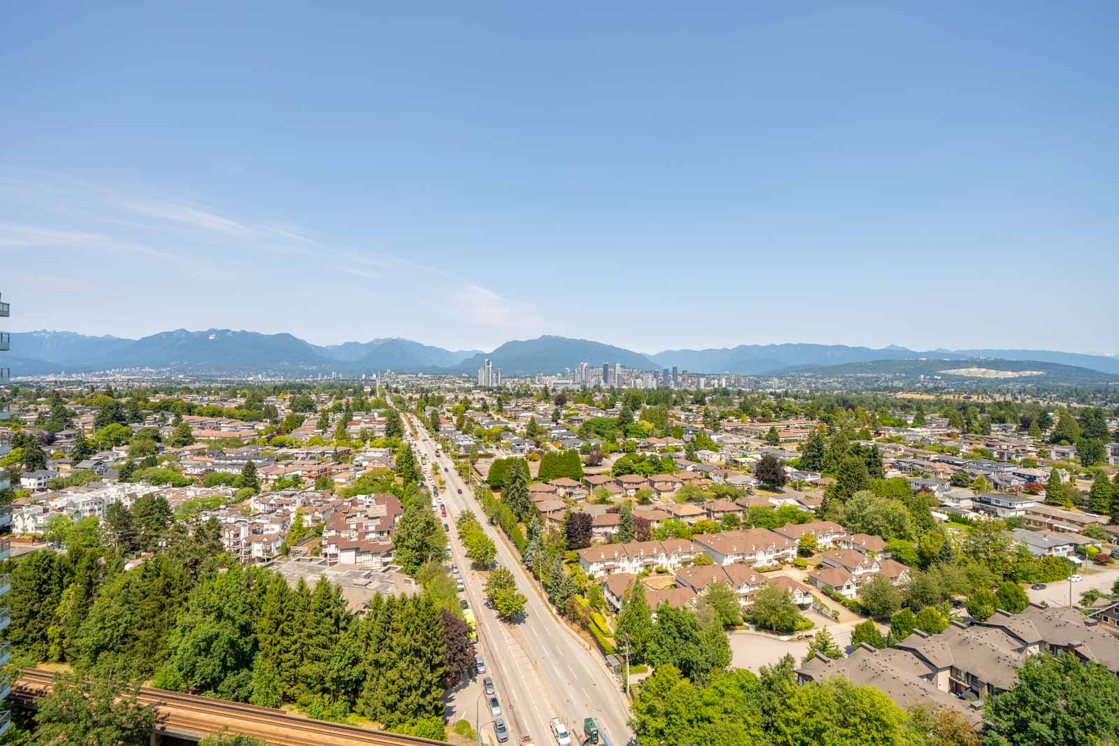 Aerial view of a Vancouver suburb along Boundary Rd, with tree-lined streets, city skyline, and mountains beneath a clear blue sky.