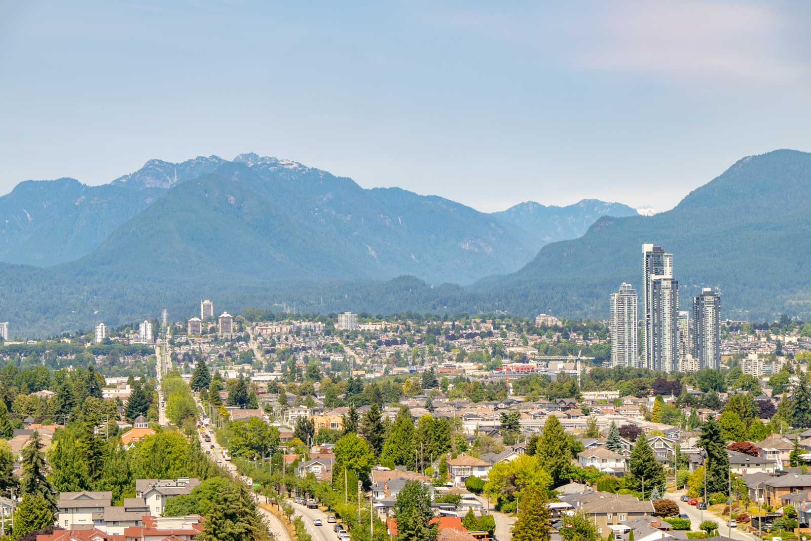 Vancouver cityscape with residential blocks, modern towers, and tree-lined Boundary Rd, backed by forested mountains and a clear sky.