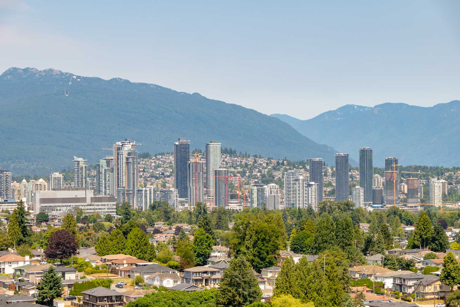 Vancouver city skyline with high-rises, tree-lined homes near Boundary Rd, and forested mountains under a clear sky.