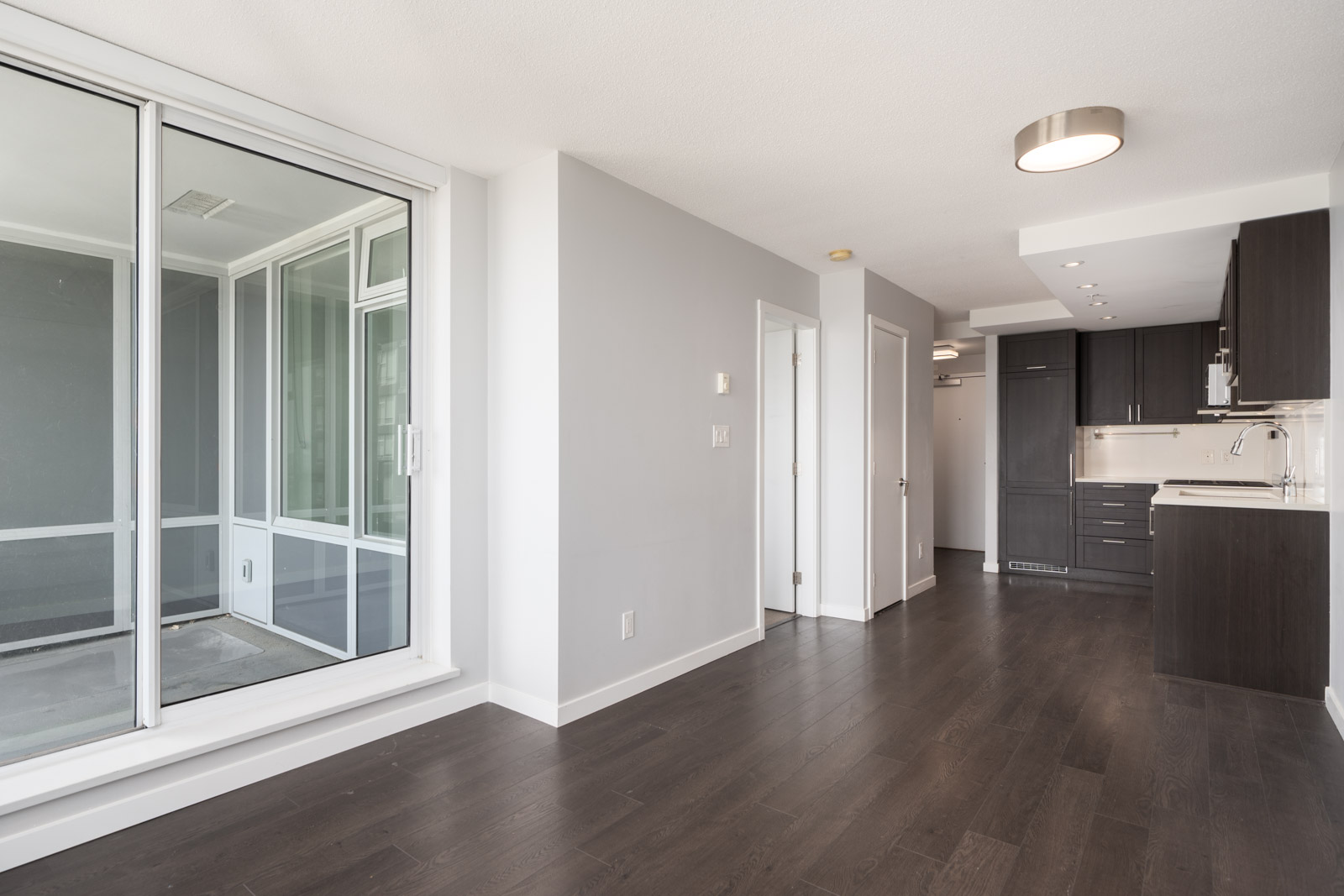 Vancouver apartment with dark wood floors, white walls, sliding balcony door, and compact kitchen with dark cabinets.