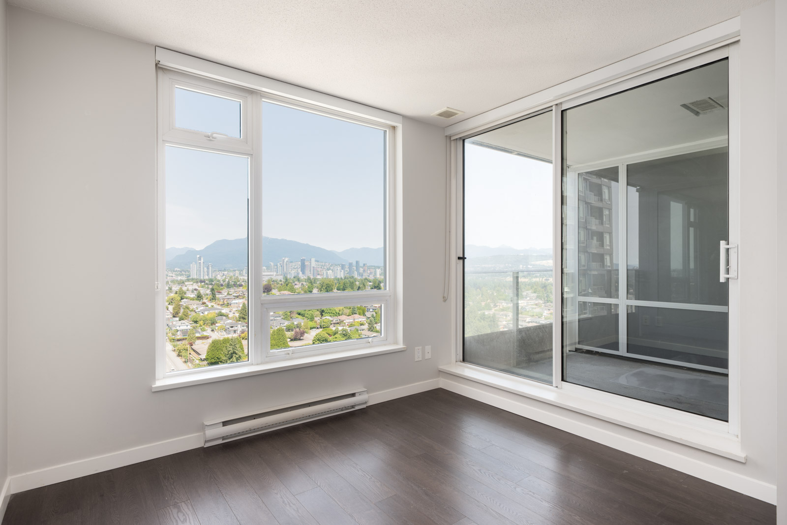 Empty modern room with dark wood floors, large window, sliding door, and balcony overlooking Vancouver city and distant mountains.