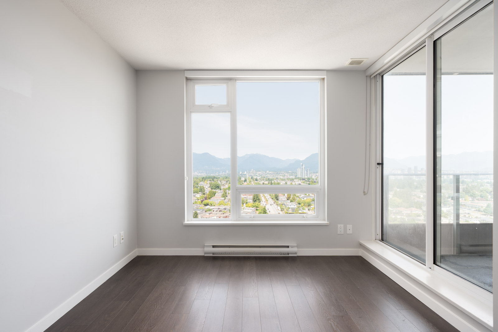 Unfurnished room with white walls, dark wood floors, large window with city and mountain views, and balcony door on Boundary Rd, Vancouver.