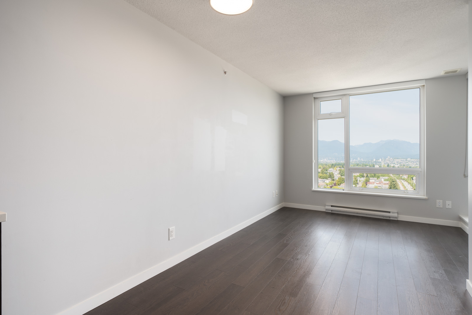 Empty room with white walls, dark wood floor, large window showing city and mountain views along Boundary Rd, Vancouver; heater below.