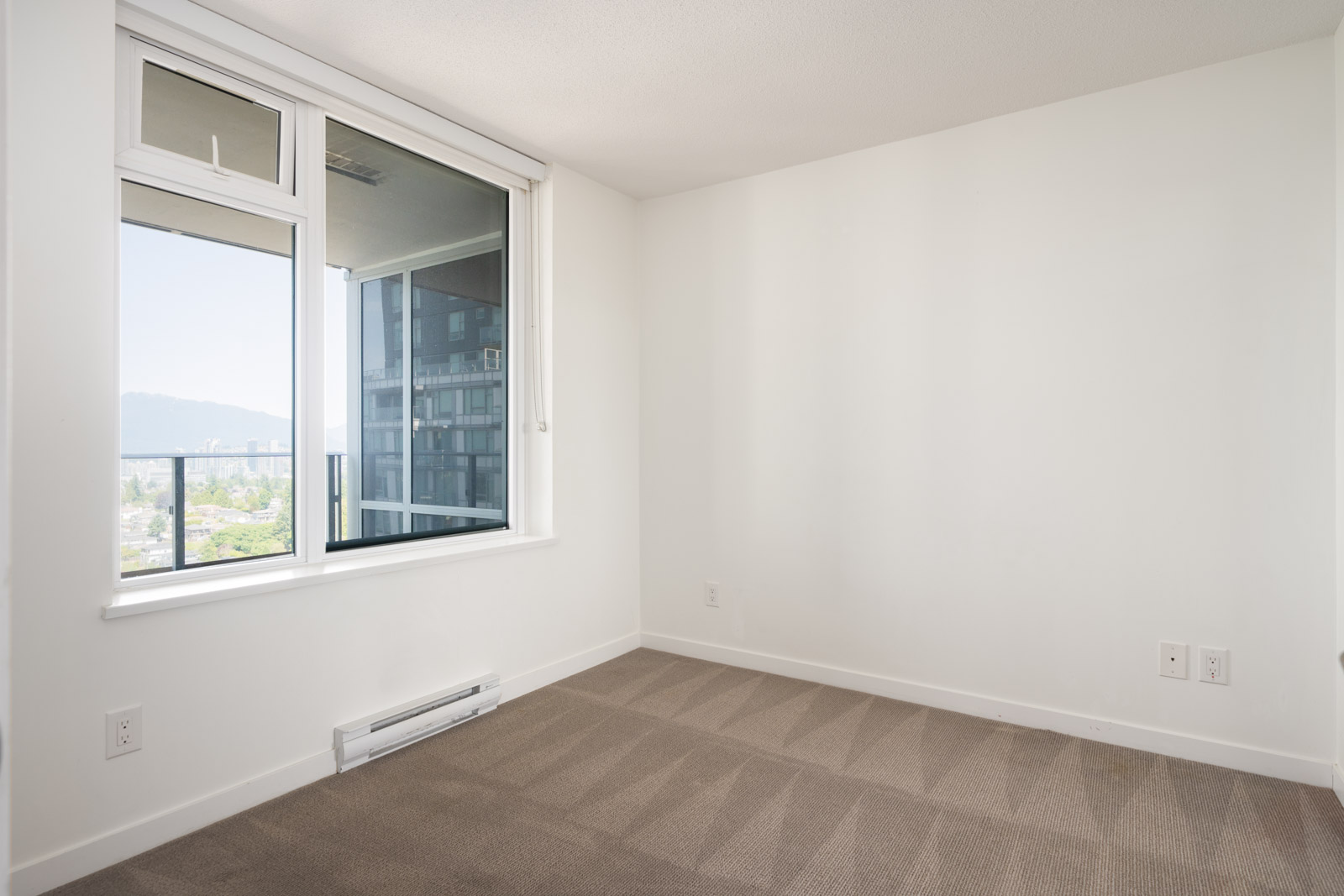 Empty room with beige carpet, white walls, large window with city view from 9th floor, and heater below on Boundary Rd, Vancouver.