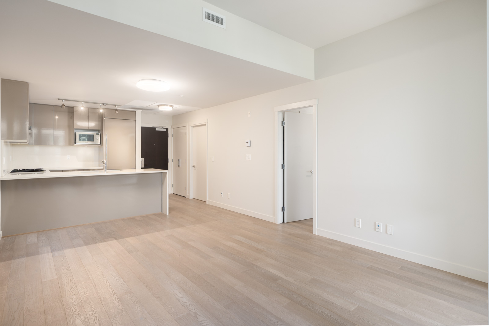 Unfurnished modern apartment with light wood flooring, white walls, a kitchen area with stainless steel appliances, and multiple doors leading to other rooms.