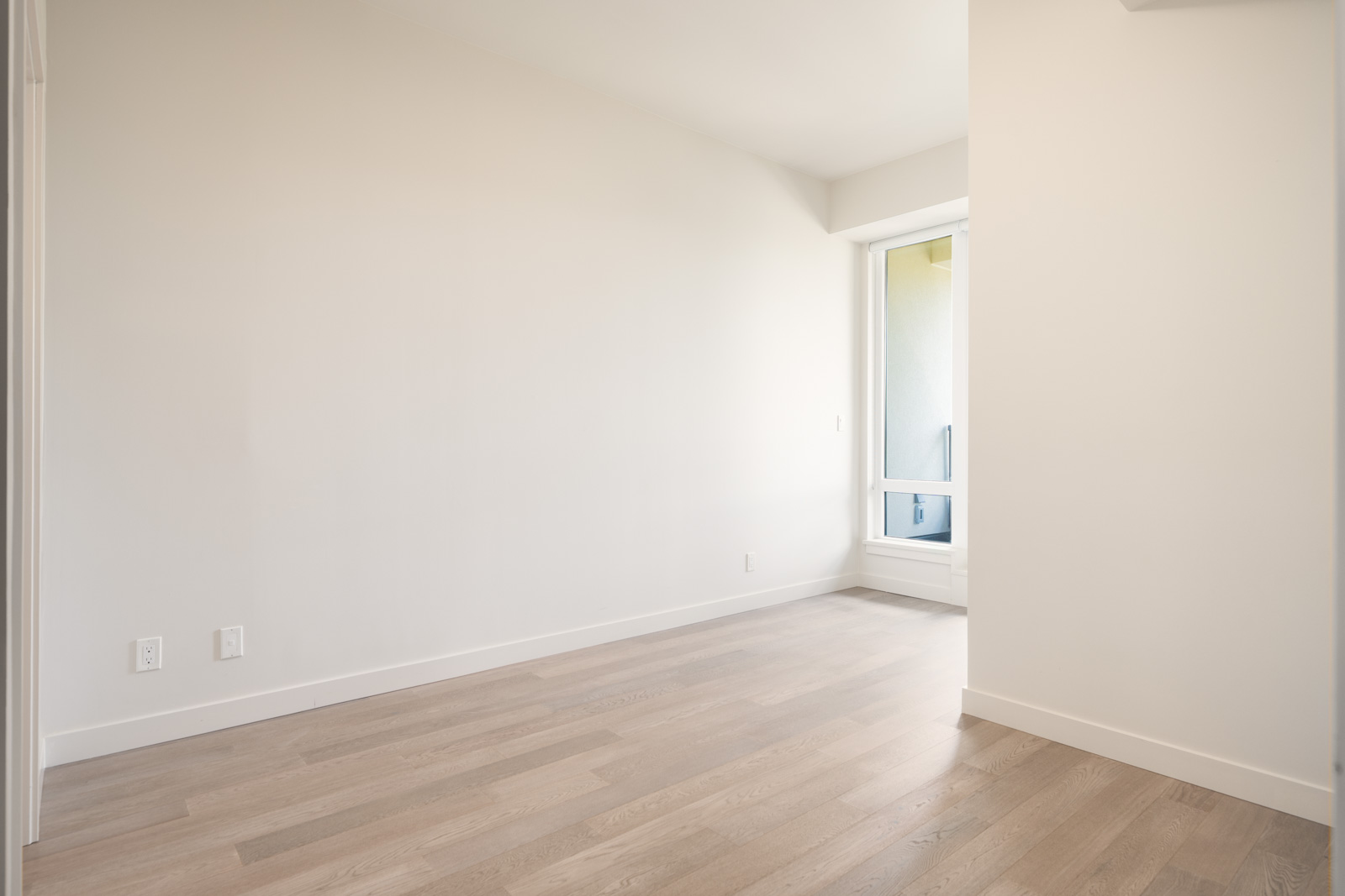 Empty room with light-colored walls, hardwood floors, and a window with a door leading to a small balcony or outdoor area.