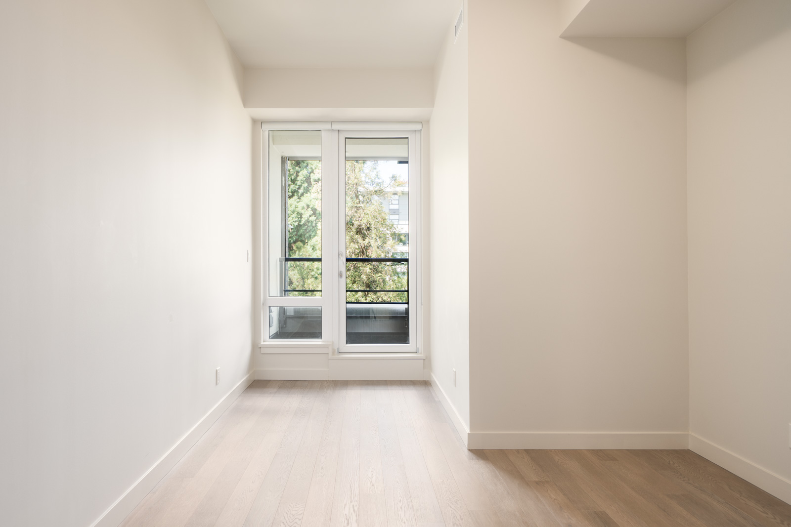 Empty room with light-colored walls and wooden floor, featuring a large window and glass door leading to a small balcony with trees visible outside.