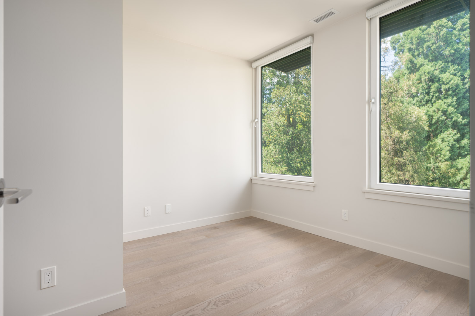Empty room with light-colored walls, large windows, and light wood flooring; trees are visible outside through the windows.