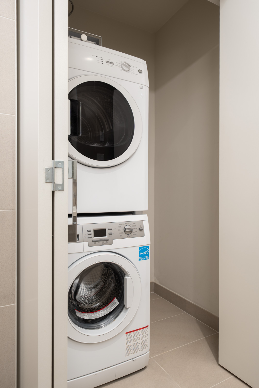 A stacked washer and dryer set is positioned in a small, tiled laundry area with neutral-colored walls and a partially open door.