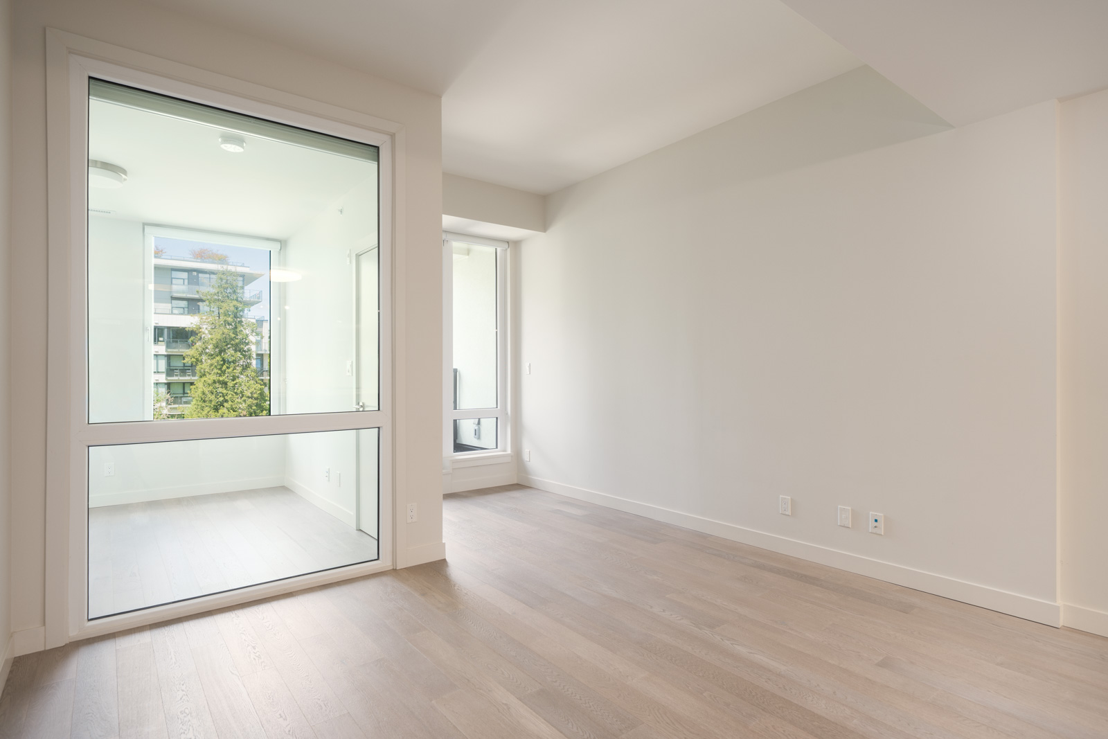 Empty modern apartment interior with light wood floors, large window, and an enclosed balcony space separated by a glass wall and door.