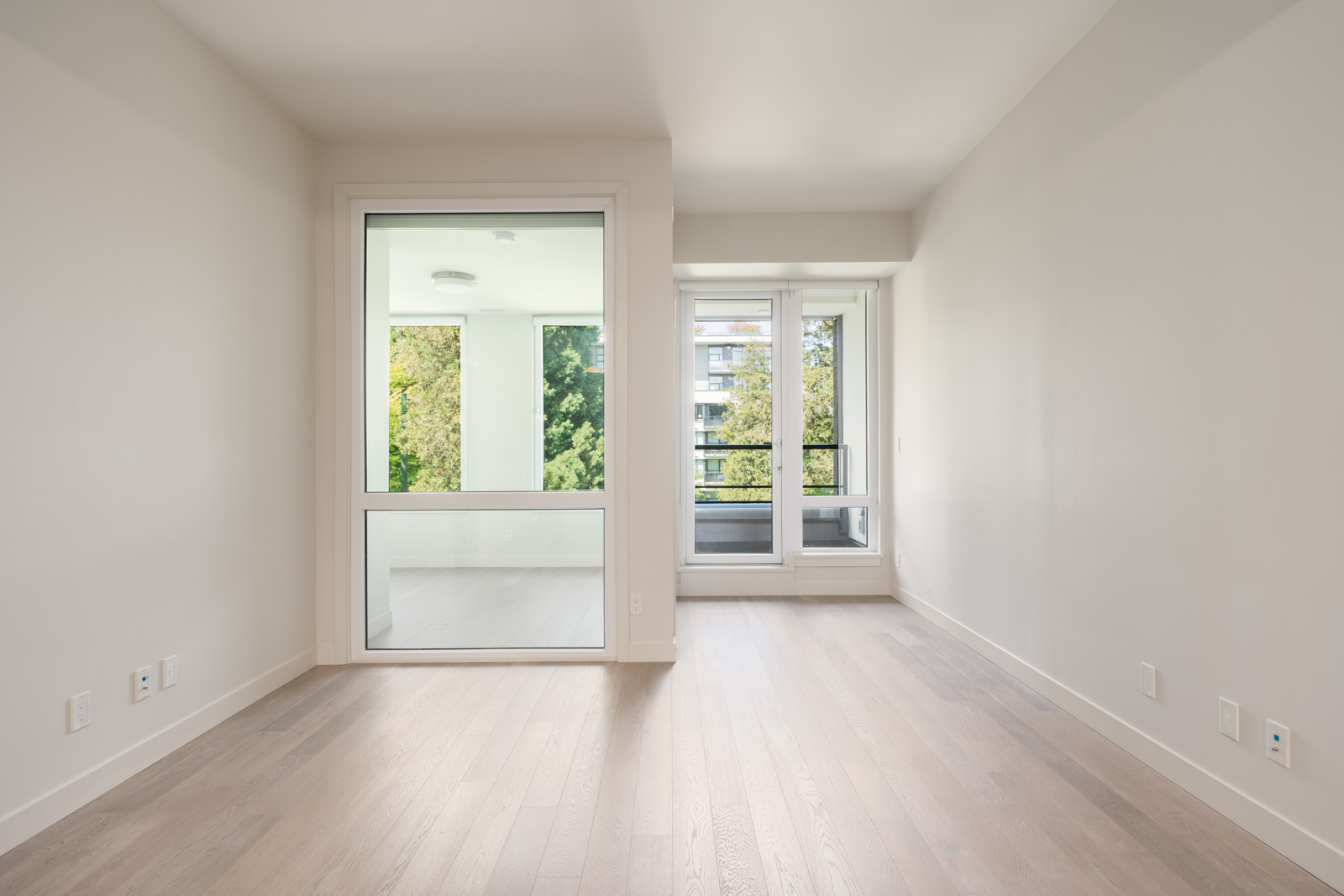 An empty, unfurnished room with light-colored wooden floors, white walls, a large interior window, and a glass balcony door overlooking trees and an apartment building.