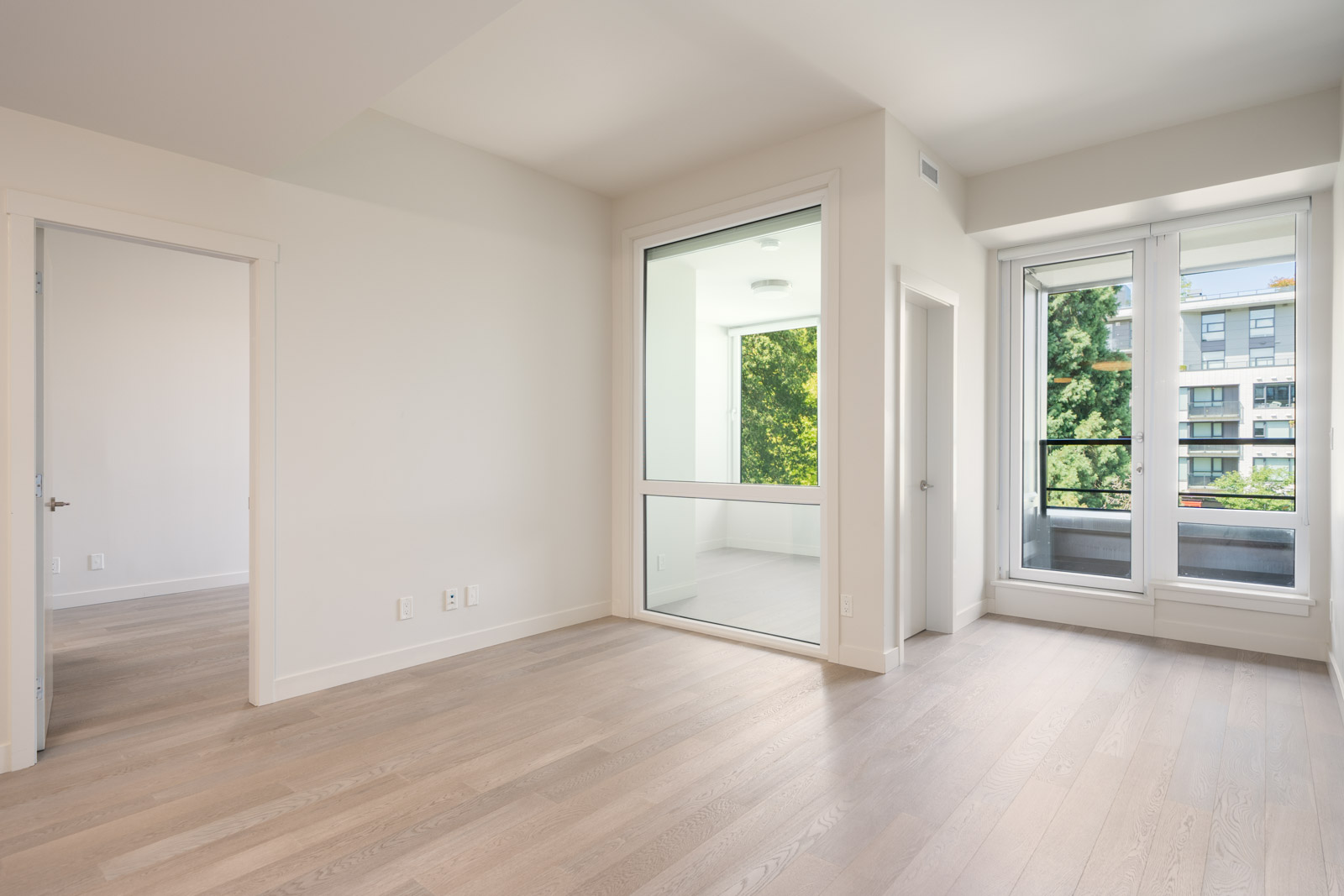 Unfurnished modern apartment room with light wood flooring, large windows, a glass-enclosed area, and a door leading to a balcony with greenery outside.