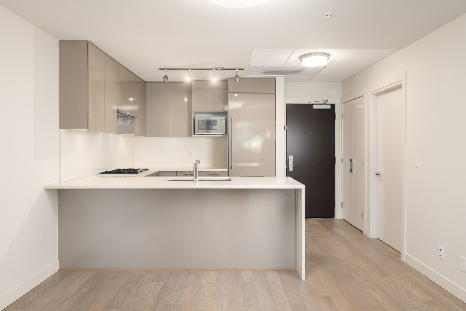 Modern apartment kitchen with beige cabinets, built-in appliances, white countertops, and a gray island, featuring light wood flooring and a dark entry door.