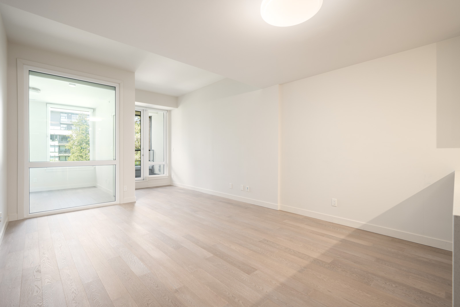 Empty room with light wood flooring, white walls, large window, and glass door leading to an enclosed balcony with outdoor view.