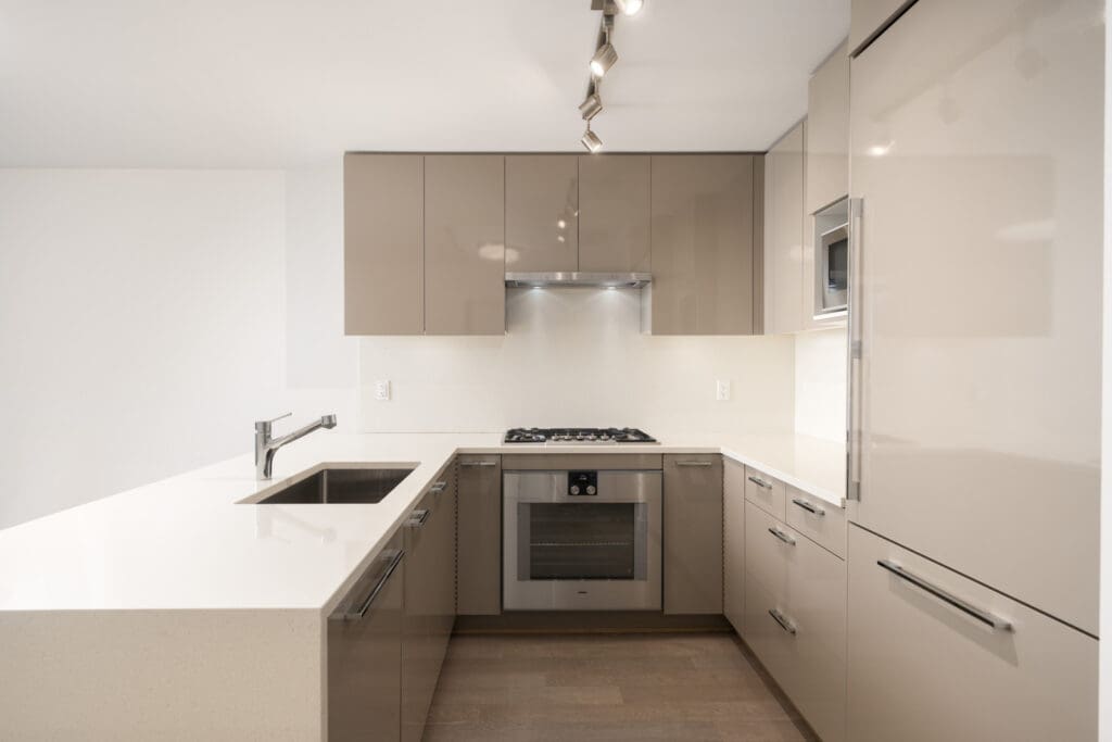 Modern kitchen with glossy beige cabinets, built-in oven, gas cooktop, sink, and overhead lighting, featuring a minimalist design and light-colored countertops.