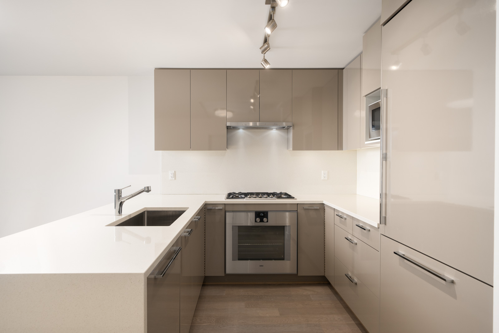 Modern kitchen with glossy beige cabinets, built-in oven, gas cooktop, sink, and overhead lighting, featuring a minimalist design and light-colored countertops.