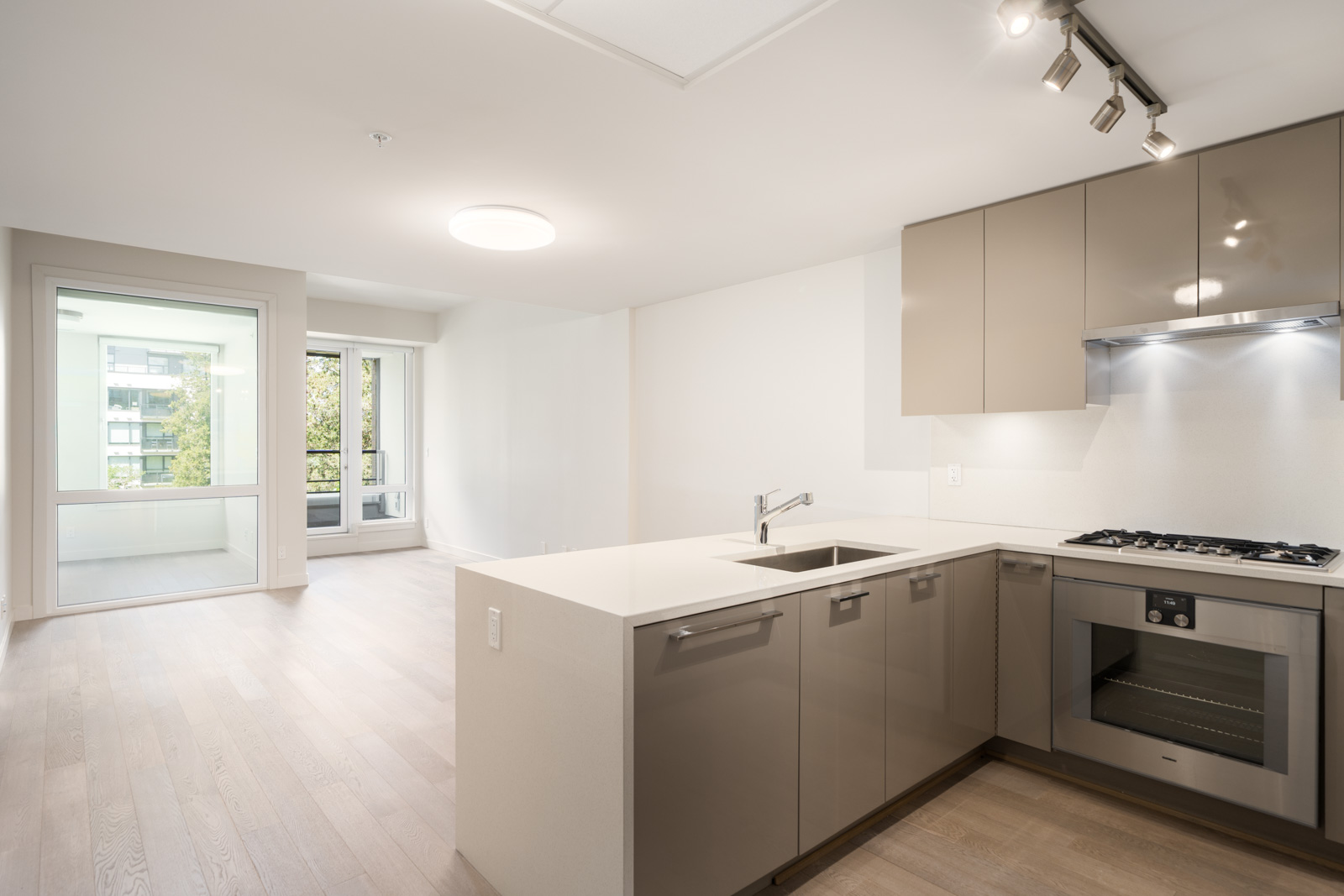 Modern kitchen with beige cabinets, stainless steel appliances, and an island sink, opening to a bright living area with large windows and light wood flooring.