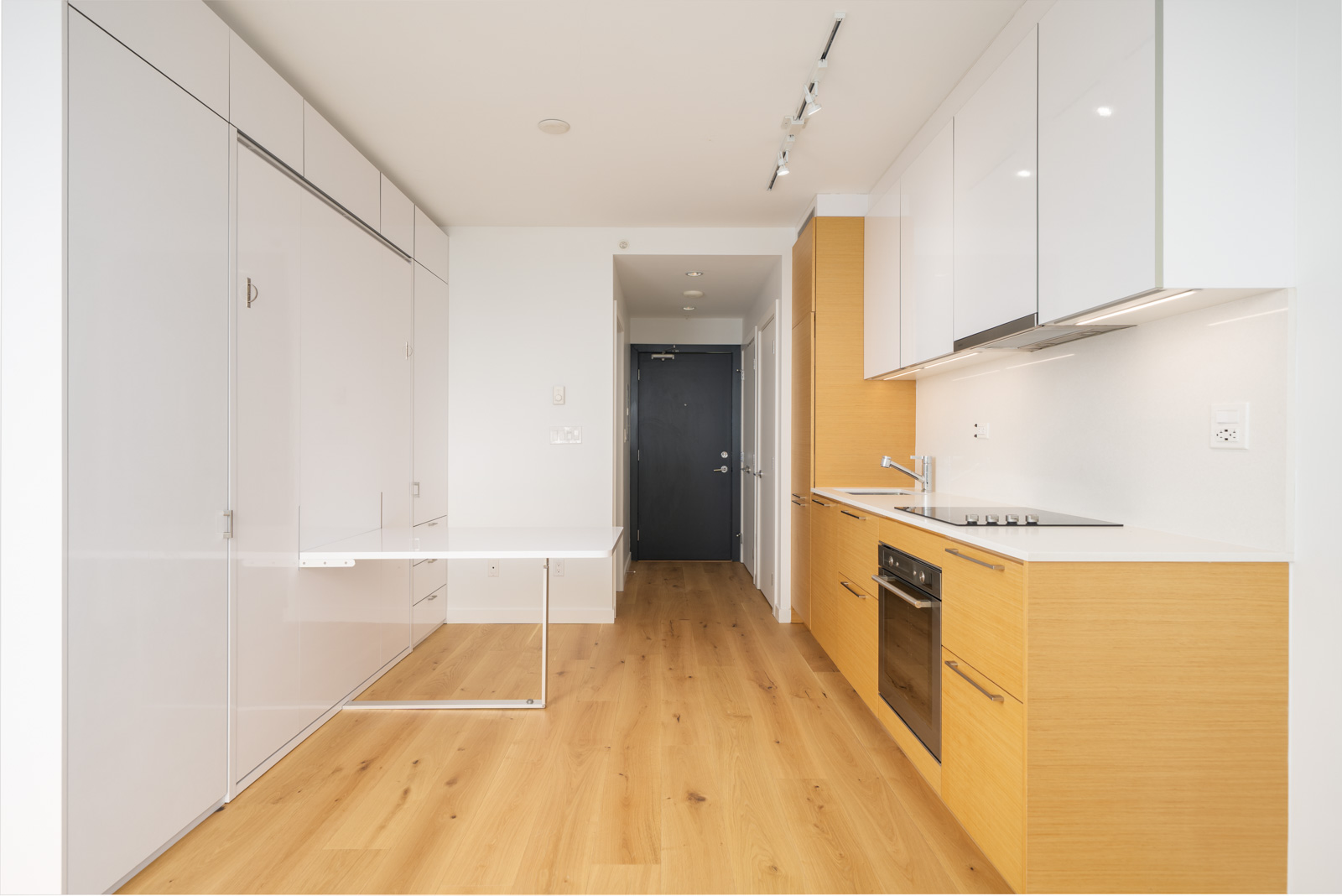 Modern minimalist kitchen with white and wood cabinets, built-in appliances, a folding wall-mounted table, and light wood flooring.